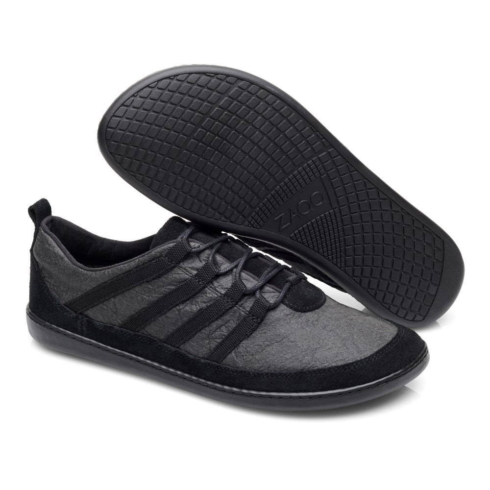 Ein Paar SPARQ Low Pinatex Black-Schuhe von ZAQQ zeigt eine minimalistische Ästhetik in Schwarz und Dunkelgrau. Ein Sneaker steht aufrecht und bietet sowohl eine Draufsicht als auch eine Seitenansicht, während der andere umgedreht ist und seine Sohle zeigt, die aus rutschhemmendem Kautschuk für verbesserte Traktion gefertigt ist.