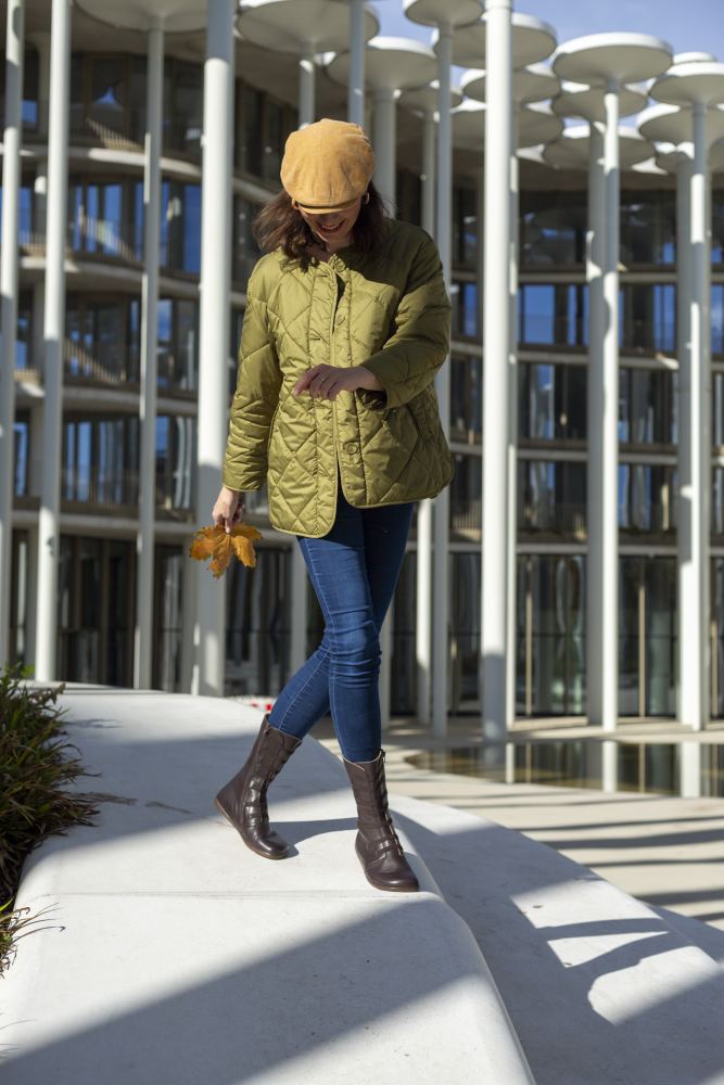 Eine Person mit gelber Mütze, grüner Steppjacke und Jeans schlendert einen weißen Weg entlang und hält mühelos die passenden ATTRAQT Stretch Nappa Brown Handschuhe von ZAQQ in den Händen. Im Hintergrund ist ein modernes Gebäude mit glänzendem Glas und eleganten Säulen zu sehen.