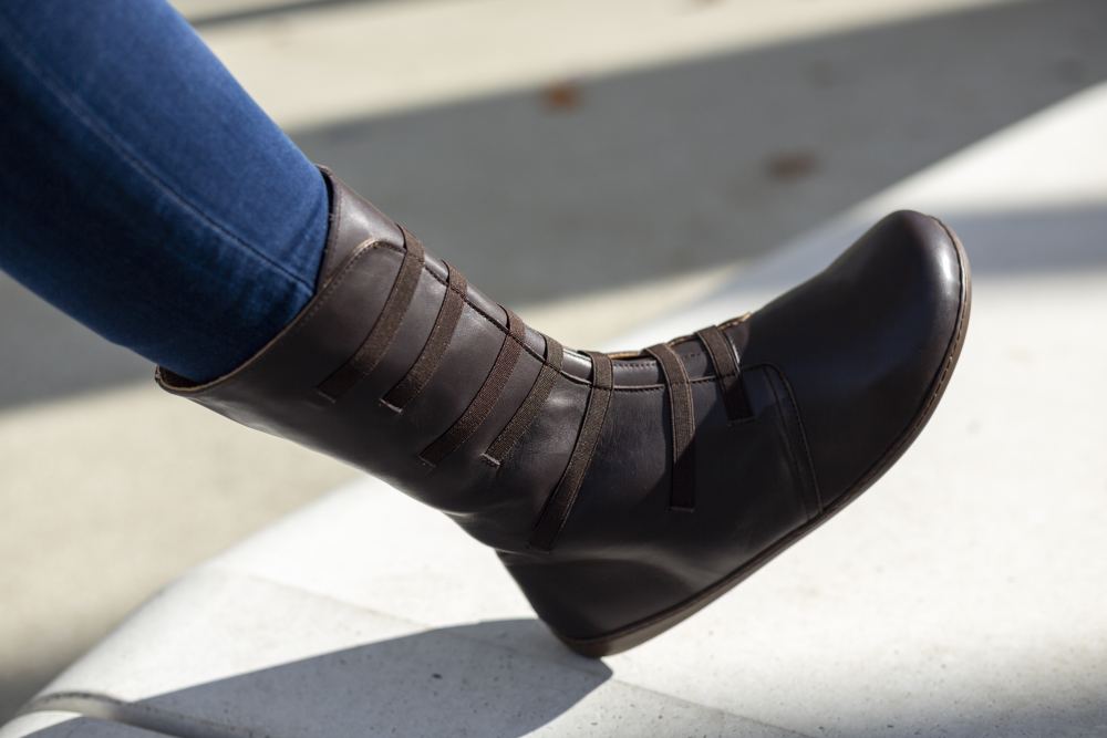 Nahaufnahme einer Person, die den knöchelhohen ATTRAQT Stretch Nappa Brown Stiefel von ZAQQ in dunkelbraunem Ziegennappa mit mehreren Riemen trägt. Kombiniert mit dunkelblauen Jeans ruht der flexible Fuß auf einer hellen Oberfläche und suggeriert so eine lässige Outdoor-Umgebung.