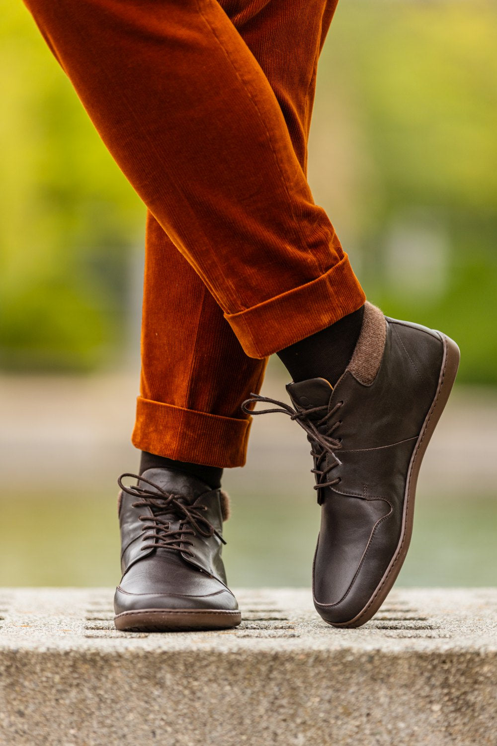 Eine Person, die Schuhe NEWQUAY Brown von ZAQQ, dazu schwarze Socken und hochgekrempelte orangefarbene Cordhosen trägt, steht auf einem Betonvorsprung. Der unscharfe Hintergrund weist Andeutungen von Grün und Gelb auf und erinnert an eine Außenkulisse.