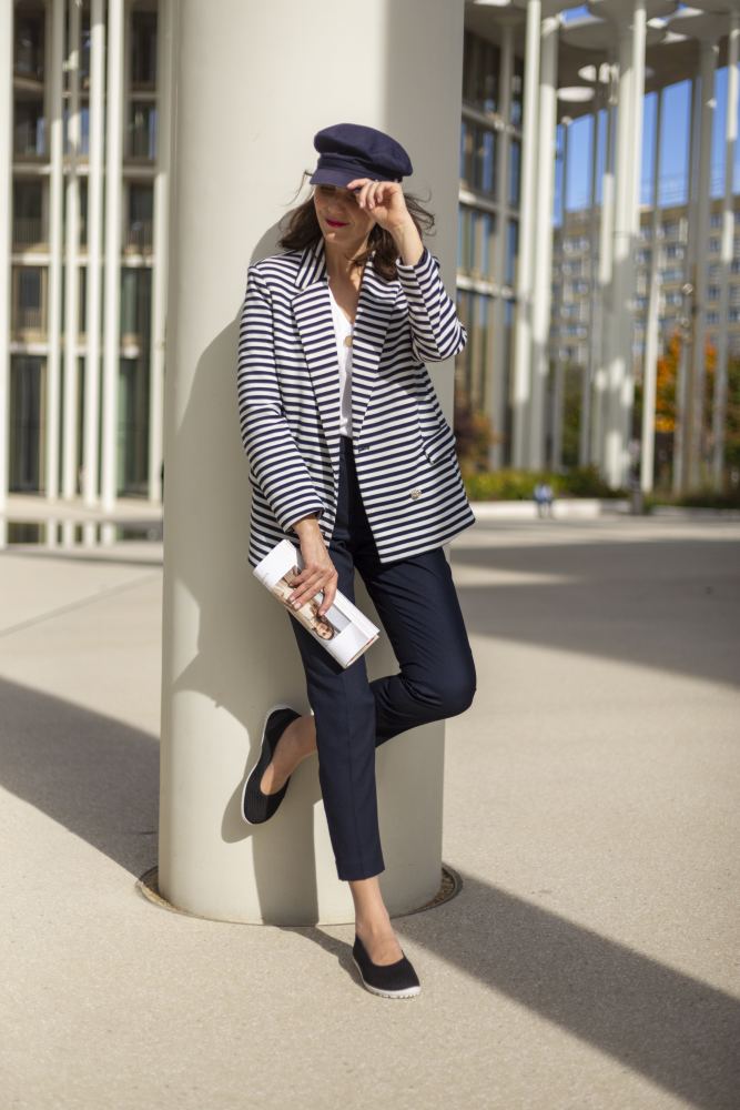 Eine Frau in gestreiftem Blazer und marineblauer Hose lehnt im Freien an einer Säule und strahlt einen femininen Look aus. Sie hält eine zusammengerollte Zeitschrift in der Hand und schützt ihre Augen mit einer marineblauen Kappe. Sonnenlicht wirft Schatten auf den Boden, wo die schwarzen SUQAR-Sneaker von ZAQQ subtile Abdrücke hinterlassen, während im Hintergrund hohe Gebäude stehen.