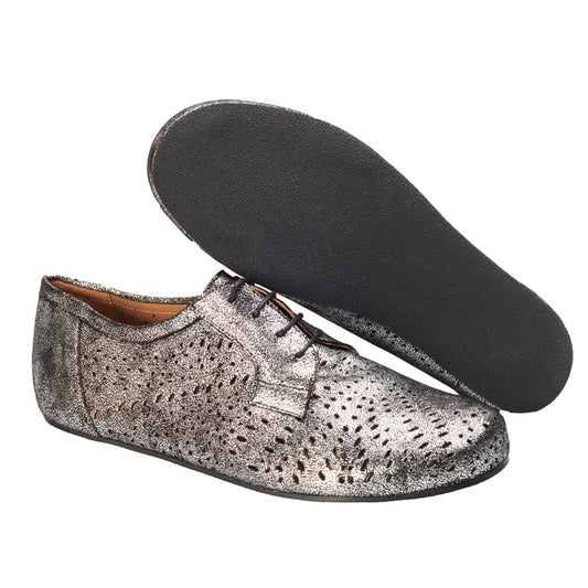 Das REMAQE Silver von ZAQQ ist ein Paar metallisch-silberner Schnürschuhe mit perforiertem Design im klassischen Derby-Schnitt. Ein Schuh steht aufrecht, um die Seite und die Oberseite hervorzuheben, während der andere umgedreht ist, um seine schwarze Sohle zu präsentieren und so dem Barfußschuh-Stil eine moderne Note zu verleihen.