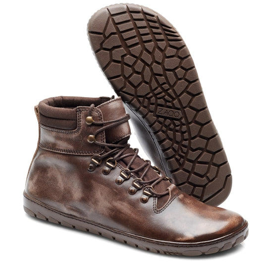 Ausgestellt ist ein Paar EXPEQ Bash Waterproof-Stiefel von ZAQQ. Diese braunen Lederschnürstiefel haben eine robuste Kautschuksohle, Metallösen und einen gepolsterten Knöchelkragen. Ein Stiefel steht aufrecht, während der andere auf der Seite liegt und das mit ZAQQ gekennzeichnete Profilmuster zeigt. Das Leder sieht leicht abgenutzt aus.