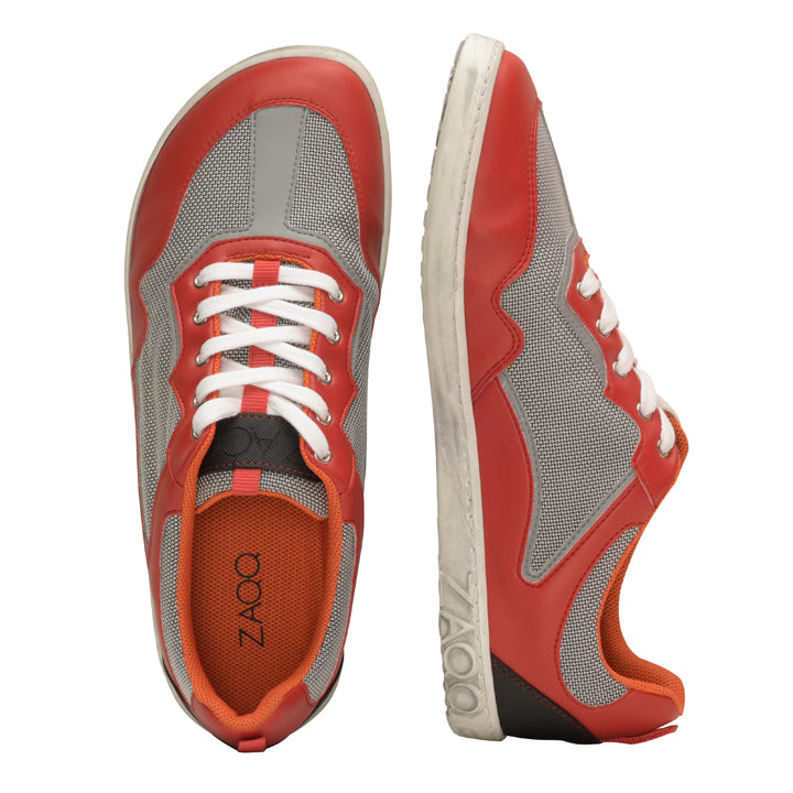 Die QOPA Red Sneaker von ZAQQ bestechen durch ihr auffälliges rot-graues Mesh-Design. Ein Sneaker steht aufrecht und hebt seine weiße Sohle mit dem ZAQQ-Logo hervor, während der andere flach liegt und sein orangefarbenes Inneres und die weißen Schnürsenkel zur Schau stellt. Diese Barfußschuhe kombinieren Stil mit Atmungsaktivität für ganztägigen Komfort.