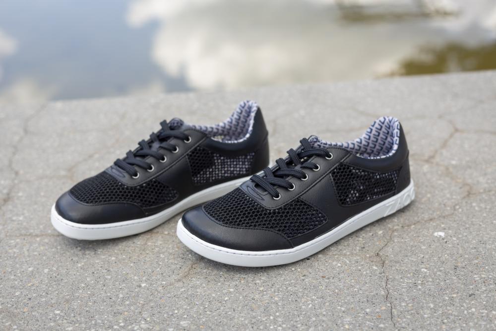 Die schwarzen ZAQQ ALIQANTE-Sneaker im Mesh-Design werden auf einer Betonoberfläche präsentiert, wobei ihr elegantes Erscheinungsbild durch die Spiegelung der Wolken im nahe gelegenen Wasser noch verstärkt wird. Diese leichteren Sneakers vereinen Stil und Komfort nahtlos.