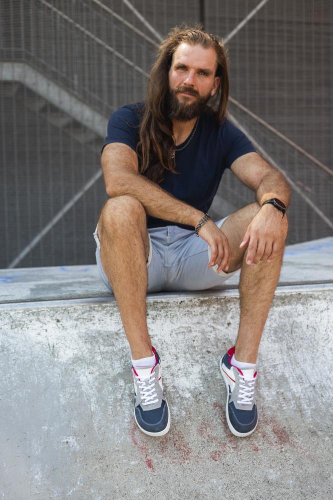 Ein bärtiger Mann mit langen Haaren sitzt auf einem Betonvorsprung, bekleidet mit einem marineblauen T-Shirt und hellgrauen Shorts. Seine BREAQ Darkblue-Sneaker von ZAQQ verleihen ihm mit ihren schwarz-weiß-roten Details Flair. Die Szene wird von einem Metallzaun und Betontreppen eingerahmt.