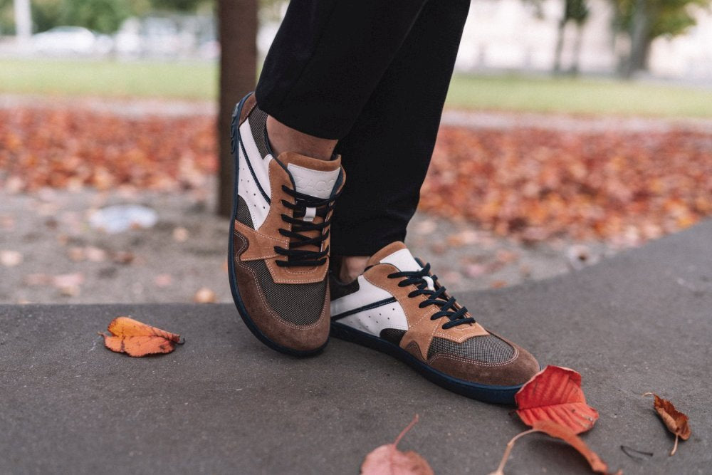 Eine Person in den schicken BREAQ Sierra Sneakers von ZAQQ mit flexibler Sohle steht auf einem Betonvorsprung. Der Boden ist mit roten und orangen Laubblättern bedeckt, die für eine herbstliche Atmosphäre sorgen.