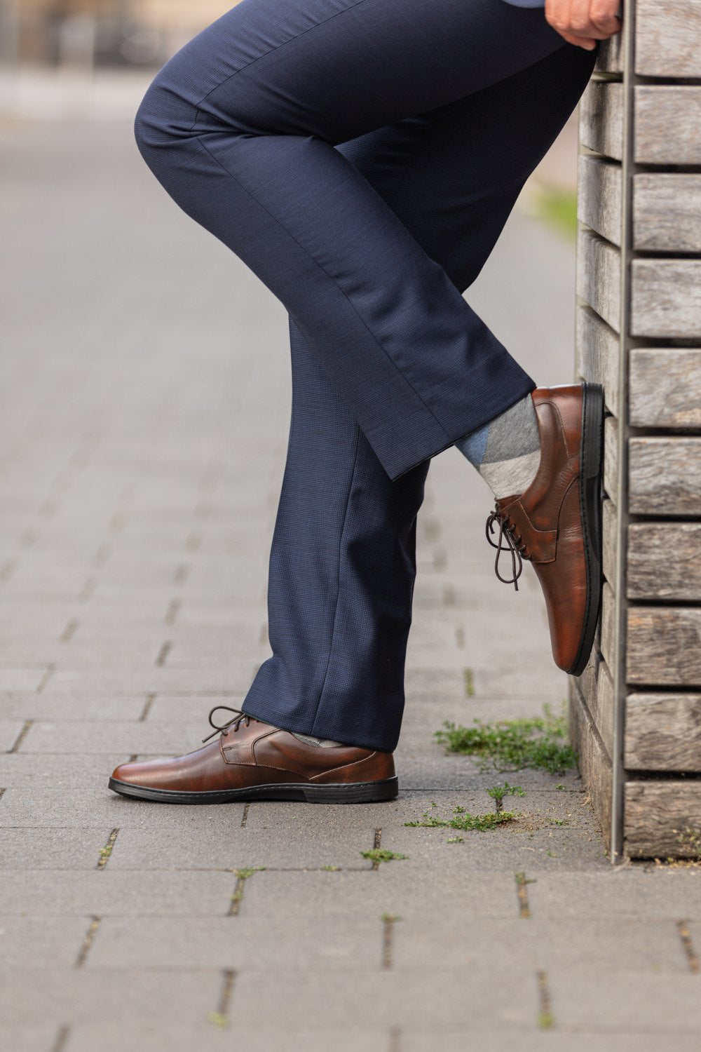 Eine Person in marineblauen Hosen und BRIQ Antique Brown-Schuhen von ZAQQ lehnt an einer Holzwand auf einem Ziegelsteinpflaster. Ein Bein ist über das andere gekreuzt, die Hände ruhen auf der Wand. Zwischen den Ziegelsteinen wachsen kleine Grasflecken.