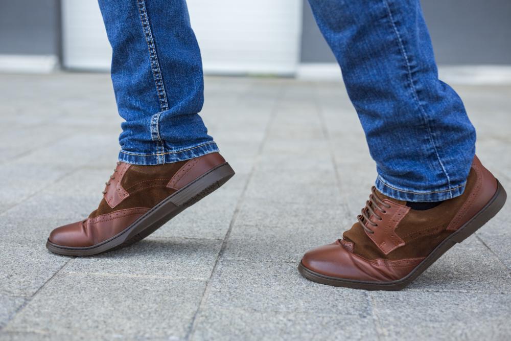 Eine Person schreitet in einem Paar BRIQ Brogue Antique Cognac-Schuhen von ZAQQ, gefertigt aus geschmeidigem Kalbsleder, über den grau gefliesten Bürgersteig. Diese dunkelbraunen Businessschuhe werden stilvoll mit leicht verwaschenen Blue Jeans kombiniert und strahlen bei jedem Schritt Raffinesse und Eleganz aus.