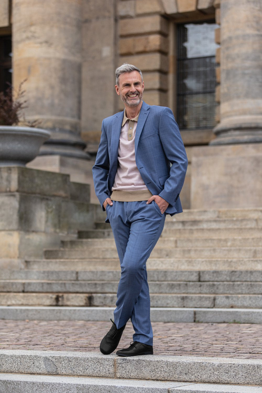 Ein Mann in blauem Anzug und hellrosa Pullover steht lässig mit den Händen in den Taschen auf Steinstufen vor einem historischen Gebäude. Er trägt die klassischen BRIQ Brogue Black-Schuhe von ZAQQ, lächelt und wirkt selbstbewusst.