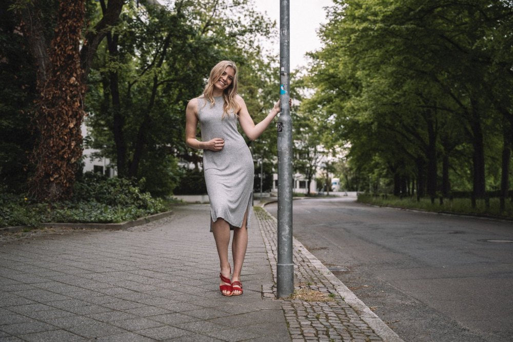 Eine Frau in einem grauen Kleid und roten Schuhen von ZAQQ lehnt an einem Laternenpfahl in einer ruhigen, von Bäumen gesäumten Straße. Sie lächelt und hat ihre rechte Hand auf dem Laternenpfahl abgestützt. Im Hintergrund sind Grünflächen und leere Bürgersteige zu sehen. In ihren stylischen Schuhen aus Veloursleder vermittelt sie ein Gefühl von heiterem Tragekomfort.