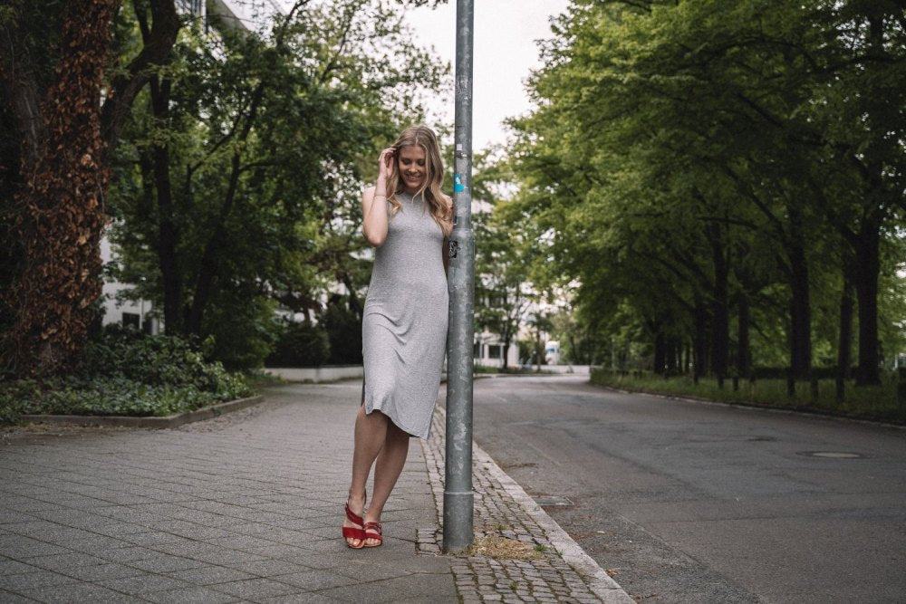 Eine Frau in einem grauen Kleid und den roten Sandalen BROOQE von ZAQQ steht neben einem Laternenpfahl in einer ruhigen, von Bäumen gesäumten Straße. Lächelnd hält sie ihr Haar, mit üppigen grünen Bäumen und einem Bürgersteig im Hintergrund, und genießt den ultimativen Komfort.