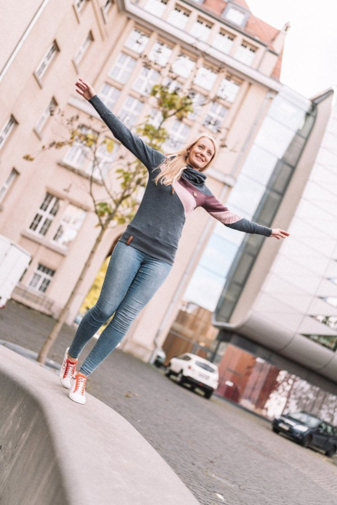 Eine Frau im lässigen Outfit, bekleidet mit Pullover und Jeans, balanciert mühelos mit ausgestreckten Armen auf einer niedrigen Betonmauer und trägt auffällige rote ZAQQ CEDRIQ-Sneaker mit Kautschuk-Sohlen. Sie befindet sich im Freien in einer städtischen Umgebung mit modernen und historischen Gebäuden im Hintergrund.