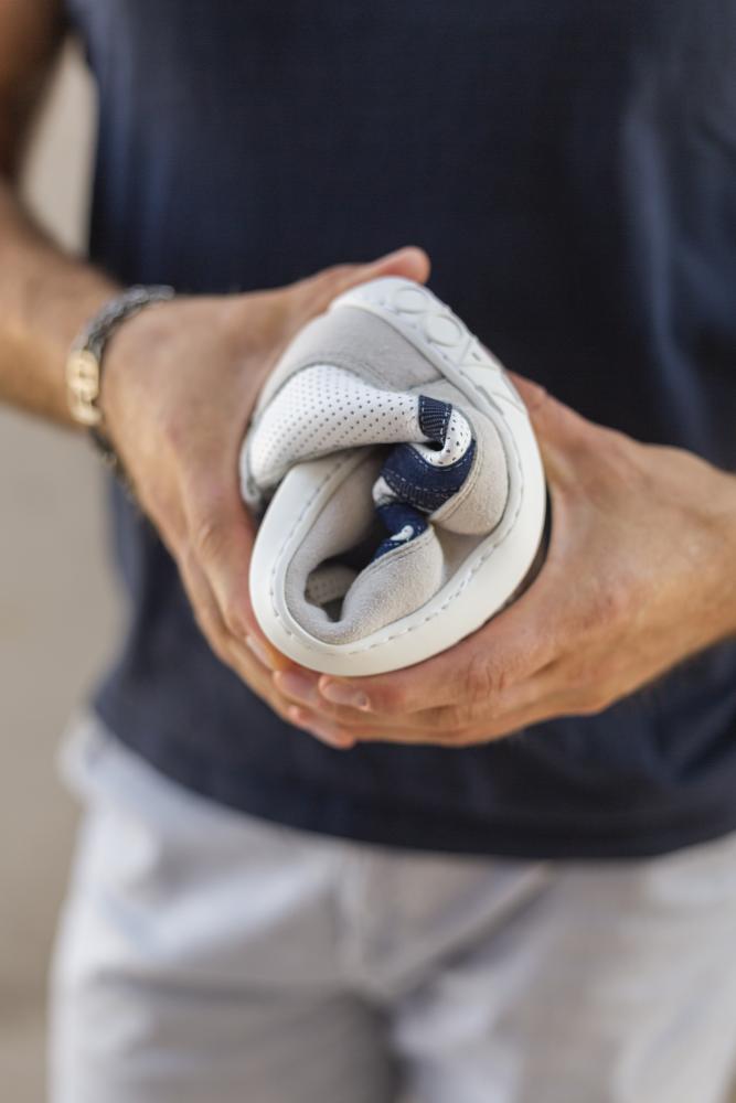 Eine Person hält einen ZAQQ DRAQE weiß-grauen Sneaker und biegt ihn, um seine Flexibilität hervorzuheben. Dieser Barfußschuh weist blaue und weiße Akzente mit Perforationen für verbesserte Atmungsaktivität auf. Die Person trägt ein dunkles Hemd und ein Armband.