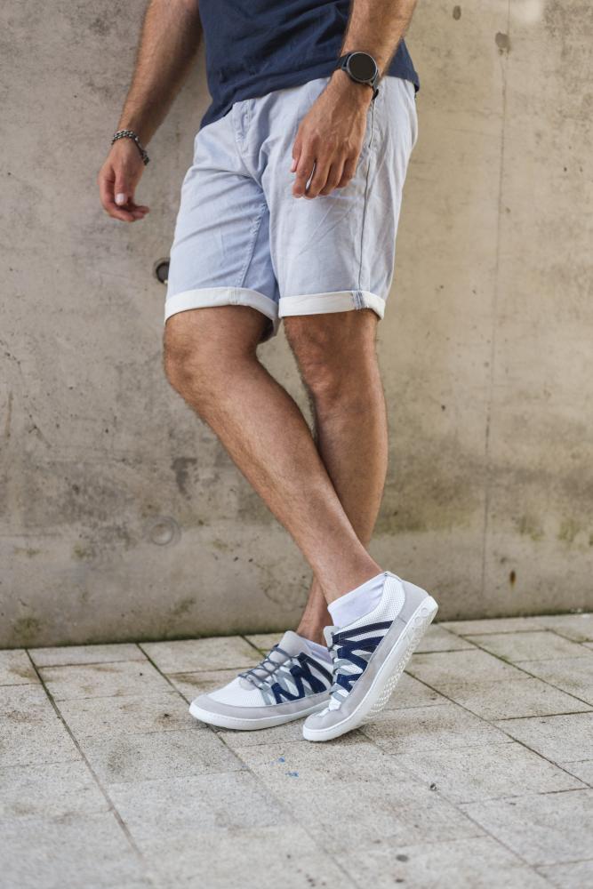 Eine Person in hellblauen Shorts und dunklem T-Shirt steht vor einer Betonwand. Sie trägt ZAQQs DRAQE White Grey Sneakers mit blauen Akzenten und hat eine Smartwatch am Handgelenk. Ihr Gesicht ist auf dem Bild nicht zu sehen.