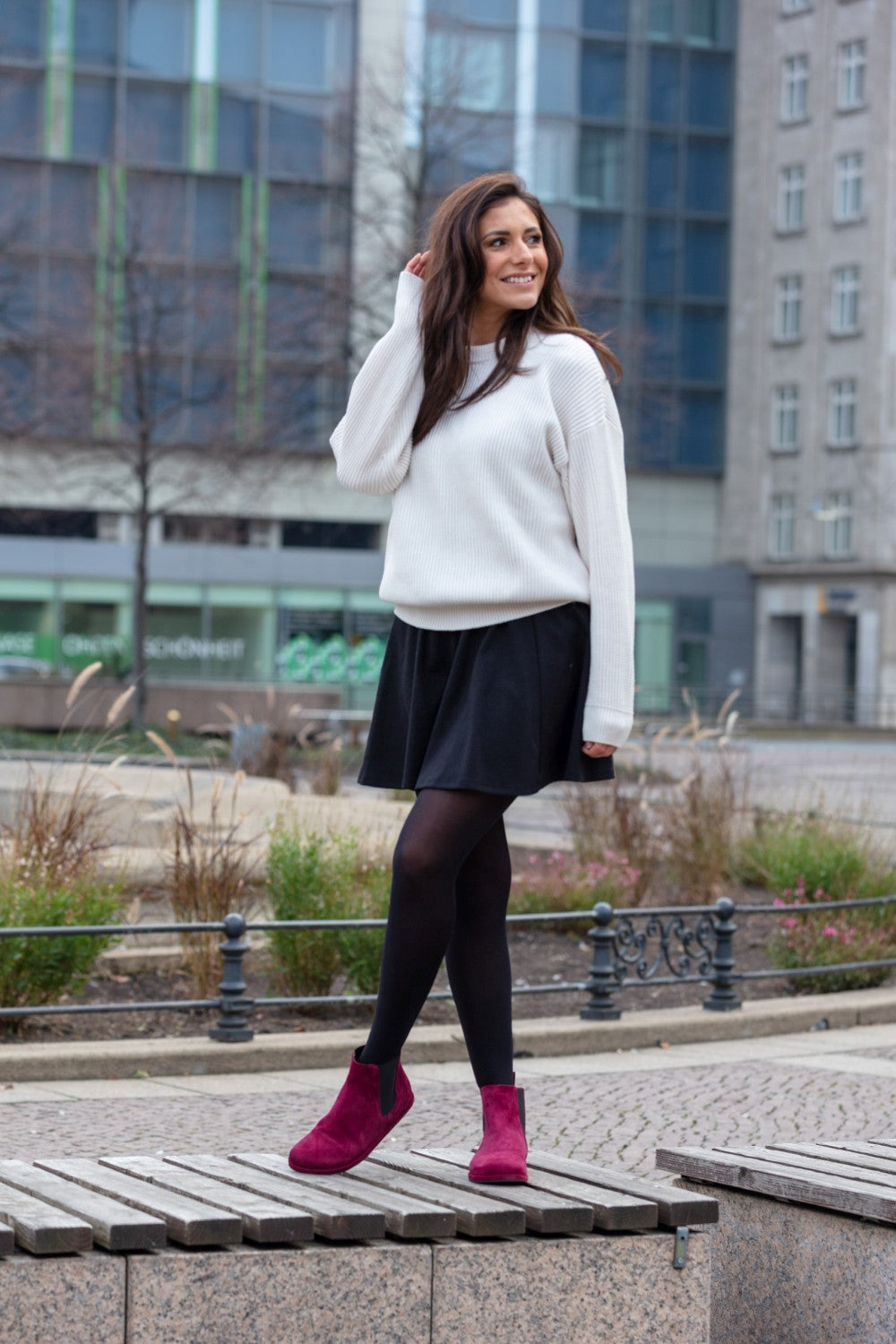 Eine Frau mit langen braunen Haaren steht auf einer Stadtstraße und trägt einen weißen Pullover, einen schwarzen Rock, schwarze Strumpfhosen und leuchtend rote ZAQQ EQUITY Berry Chelsea Boots. Sie lächelt und berührt mit einer Hand ihr Haar. Im Hintergrund sind Gebäude und Bänke zu sehen.