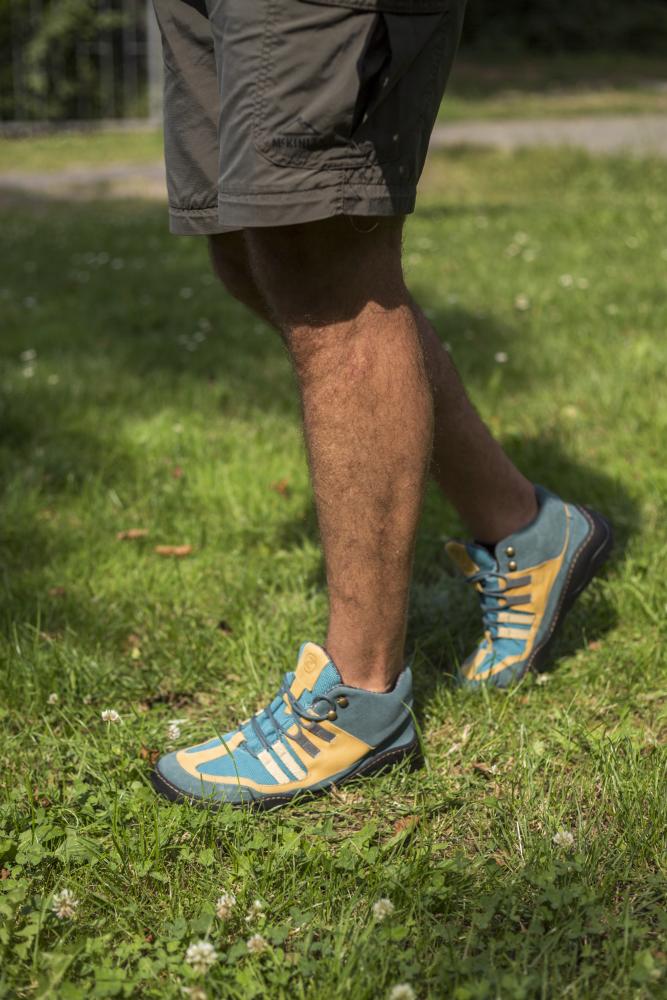 Eine Person läuft auf Gras. Sie trägt graue Shorts und die ZAQQ ESQAPE Blue Orange Waterproof Barfuß-Sneaker. Das grüne Gras ist mit kleinen weißen Blumen übersät und im Hintergrund ist ein Pfad zu sehen.