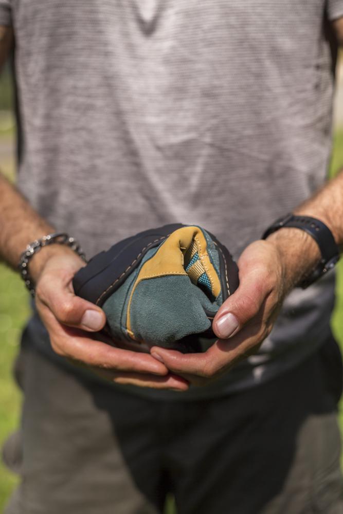 Eine Person mit gestreiftem Hemd und Armbändern hält mit beiden Händen einen kleinen, bunten, weichen Beutel. Dieser Beutel mit seinen lebendigen, an die Natur erinnernden Farbtönen zeigt Farben wie Blaugrün, Gelb und Schwarz vor einem Hintergrund aus verschwommenem Grün. Sein nahtloses Design ähnelt der Handwerkskunst des ZAQQ ESQAPE Blue Orange Waterproof.