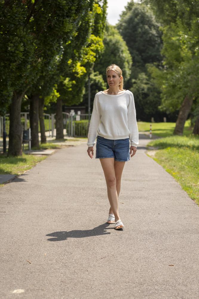 Eine Frau in einem weißen ZAQQ FLAQE Pullover und Jeansshorts geht an einem sonnigen Tag einen von Bäumen gesäumten Weg entlang, ihre Schritte sind anmutig in Barfuß-Sandalen aus geschmeidigem Nappaleder.