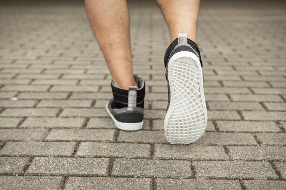 Nahaufnahme einer Person, die auf einem Steinpflaster läuft. Hervorgehoben wird die Rückansicht der stylischen ZAQQ FRANQY Black Midcut-Sneaker. Die leicht gebeugten Beine betonen die Schuhsohlen, während sie selbstbewusst nach vorne treten.