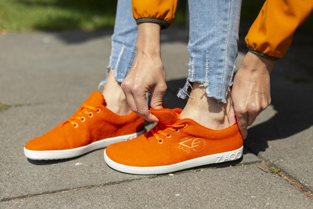 Eine Person in Jeans mit ausgefranstem Saum und orangefarbener Jacke bindet die Schnürsenkel der leuchtend orangefarbenen LIQE Orange-Sneaker von ZAQQ, die eine leuchtend orange Farbe mit weißen Sohlen haben. Diese Schuhe vermitteln ein Barfußgefühl und vereinen mühelos Stil und Komfort.