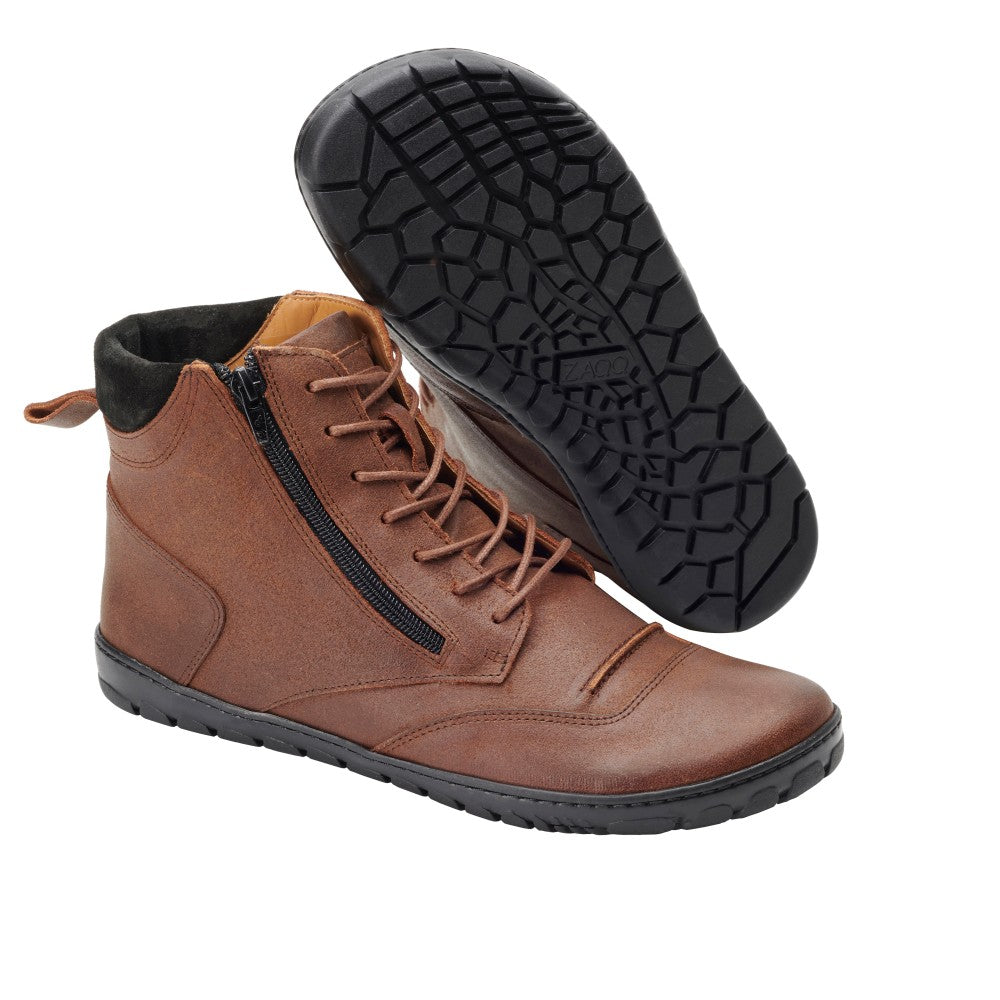 Der PARQER Sierra von ZAQQ ist ein Paar braune Lederstiefeletten mit klassischem Design und schwarzer, flexibler Sohle. Ein Stiefel steht aufrecht und hebt die Schnürsenkel und den seitlichen Reißverschluss hervor, während der andere so angeordnet ist, dass er die strukturierte Sohle zur Geltung bringt.