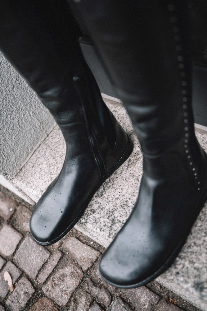 Eine Nahaufnahme der stylischen QATCH SHINE Nappa Black-Stiefel von ZAQQ, elegant verziert mit kleinen Wassertropfen auf der Oberfläche, auf einer Steinstufe stehend. Das Kopfsteinpflaster darunter lässt eine nasse Außenumgebung vermuten.