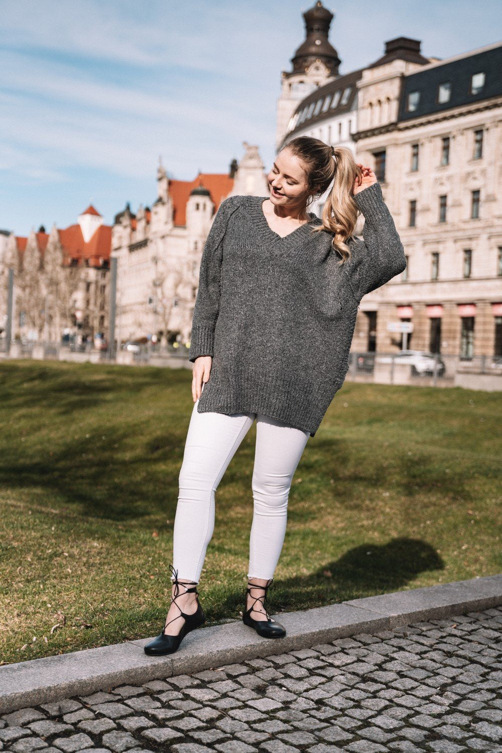 Eine Person mit langen hellen Haaren steht auf einem Kopfsteinpflasterweg und hält verspielt ihren Pferdeschwanz. Sie trägt einen grauen Pullover und eine weiße Hose, ergänzt durch stylische schwarze ZAQQ QISS Schuhe aus Nappaleder. Im Hintergrund sind eine Rasenfläche und historische Gebäude unter einem klaren blauen Himmel zu sehen.