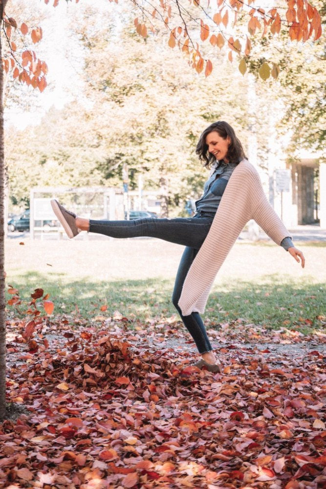 Eine Frau tritt freudig einen Haufen Herbstblätter in einem Park, ihre beige Strickjacke schwingt mit ihren Bewegungen. Sie trägt gemütliche QOMFY Wide Grey Schuhe von ZAQQ aus Veloursleder, perfekt geeignet für den sonnigen Herbsttag zwischen Bäumen, die mit orangefarbenen und gelben Blättern geschmückt sind.