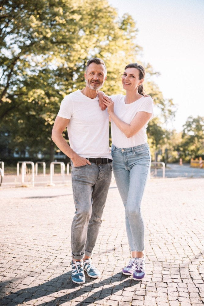 Ein Paar steht lächelnd an einem sonnigen Tag im Freien, beide tragen weiße Hemden und Jeans. Der Mann, aus dem RIQ Purple Sneakers von ZAQQ hervorschauen, hat die Hände in den Taschen. Die Frau legt ihre Hand auf seine Schulter. Bäume und ein klarer Himmel bilden den Hintergrund dieses ungezwungenen Moments.