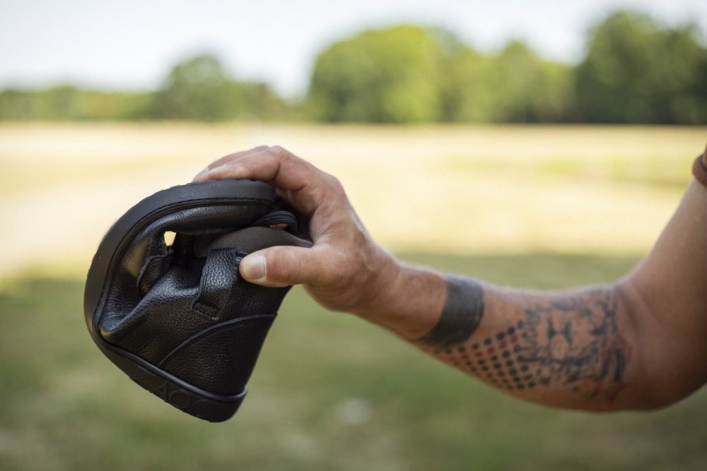 Der tätowierte Arm einer Person hält den ROQA Black Sneaker von ZAQQ und biegt ihn in der Mitte, um seine Flexibilität zu demonstrieren. Der Hintergrund zeigt eine verschwommene Außenkulisse mit Gras und Bäumen, die das elegante schwarze Erscheinungsbild des Schuhs ergänzt.