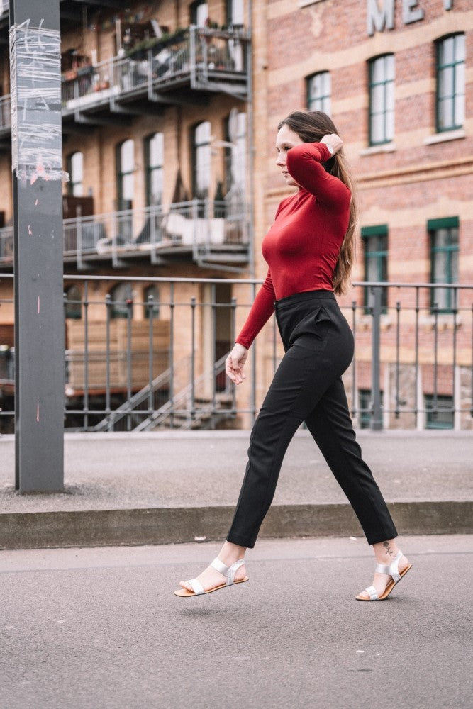 Eine Frau geht selbstbewusst eine Straße entlang. Sie trägt ein rotes Langarmshirt und eine schwarze Hose, dazu minimalistische ZAQQ SLIQ-Silver-Sandalen. Mit wehendem langen Haar schlendert sie an einem Geländer vorbei, im Hintergrund sind Backsteingebäude zu sehen.