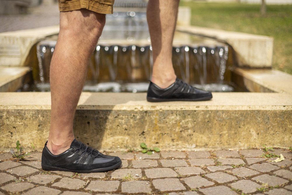 Eine Person, die die SPARQ Low Black Sneaker von ZAQQ und braune Shorts trägt, steht auf einer Steinkante vor einem fließenden Brunnen. Der Boden ist mit Kopfsteinpflaster gepflastert, im Hintergrund ist Gras zu sehen.