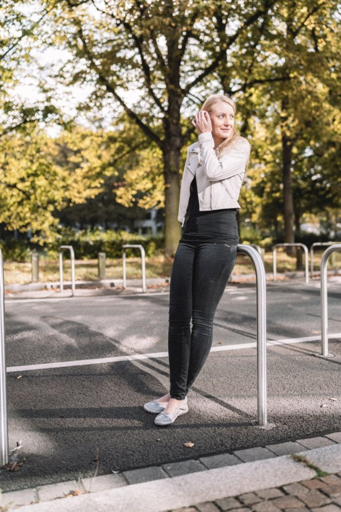 Eine Person mit langen blonden Haaren, die eine weiße Jacke und dunkle Jeans trägt, ergänzt durch ZAQQs SUQAR Grey-Schuhe, steht auf einer Straße und lehnt an einem Metallpfosten. Im Hintergrund sind Bäume mit grünen Blättern zu sehen, und Sonnenlicht fällt durch die Äste.