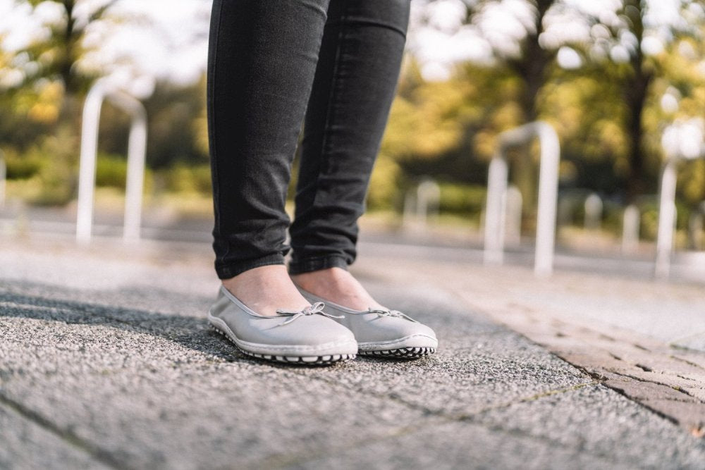 Eine Person steht im Freien auf einer gepflasterten Fläche und trägt SUQAR Grey Flats von ZAQQ und eine schwarze Hose. Im Hintergrund sind verschwommene Bäume und weiße Pfosten zu sehen, was an einen Park oder eine Fußgängerzone an einem sonnigen Tag erinnert.