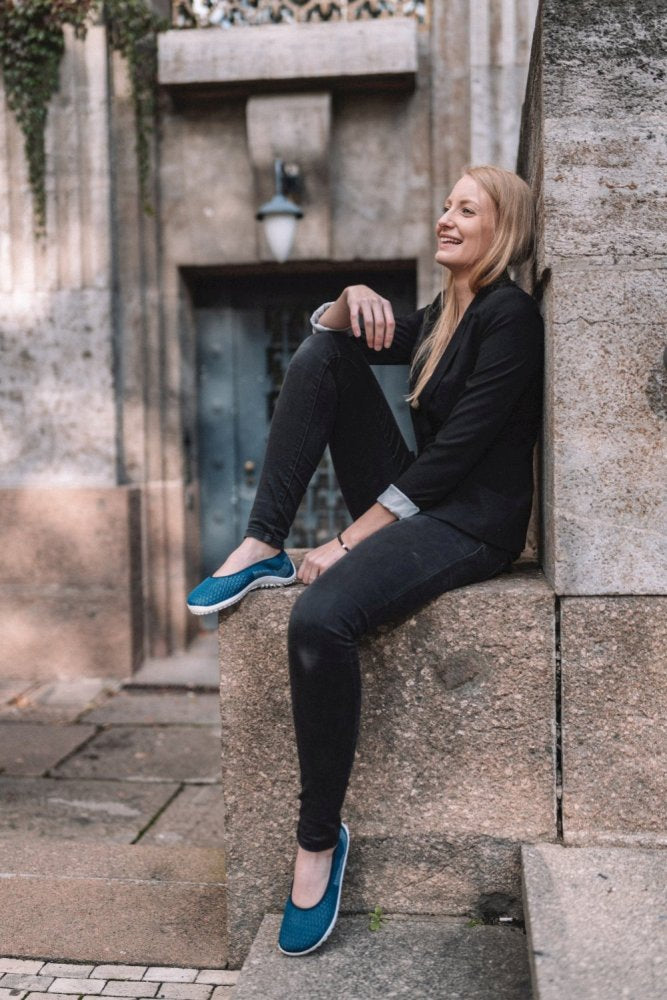 Eine Person mit langen Haaren, gekleidet in ein schwarzes Outfit und blaue SUQAR Mare-Schuhe von ZAQQ, sitzt auf einem Steinvorsprung vor einem alten Gebäude. Sie lächelt und blickt nach vorne, wobei ein Fuß auf dem Vorsprung ruht und eine Hand auf ihrem Knie liegt.