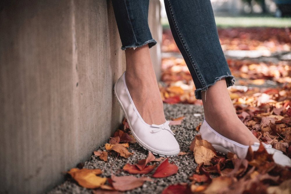 Eine Person in weißen SUQAR Ballerinas von ZAQQ und ausgefransten Jeans lehnt an einer Wand. Der Boden ist mit bunten Herbstblättern bedeckt, was für eine gemütliche Herbststimmung sorgt.