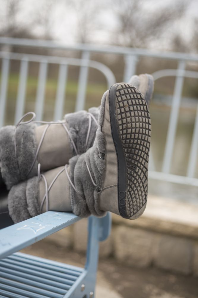 Ein Paar graue, pelzgefütterte TORQ Winterstiefel von ZAQQ stehen auf einer hellblauen Metallbank. Die Sohlen weisen ein markantes gitterartiges Muster auf, das für ein Barfußgefühl sorgt. Im Hintergrund ist ein unscharfer Park mit einem Geländer und kahlen Bäumen zu sehen.