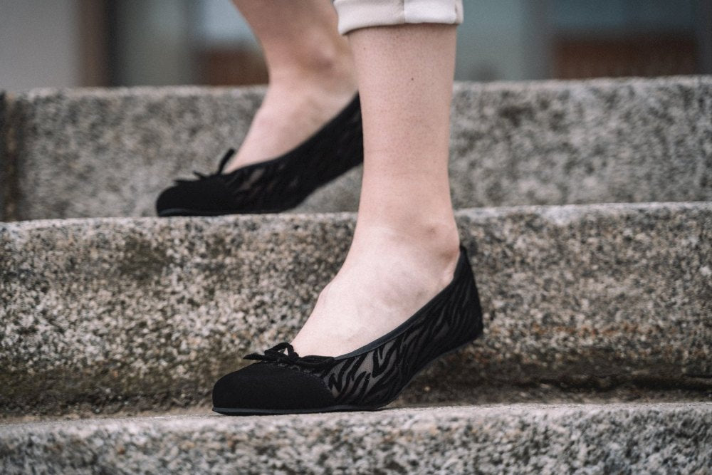 Eine Person in schwarzen Ballerinas mit dezentem Muster steht auf einer Steintreppe. Die Hose ist an den Knöcheln hochgekrempelt und die Schuhe ZAQQ TWIST Sling Black haben oben eine kleine Schleife und bieten mit ihrem flexiblen Sohlendesign sowohl Style als auch Komfort.