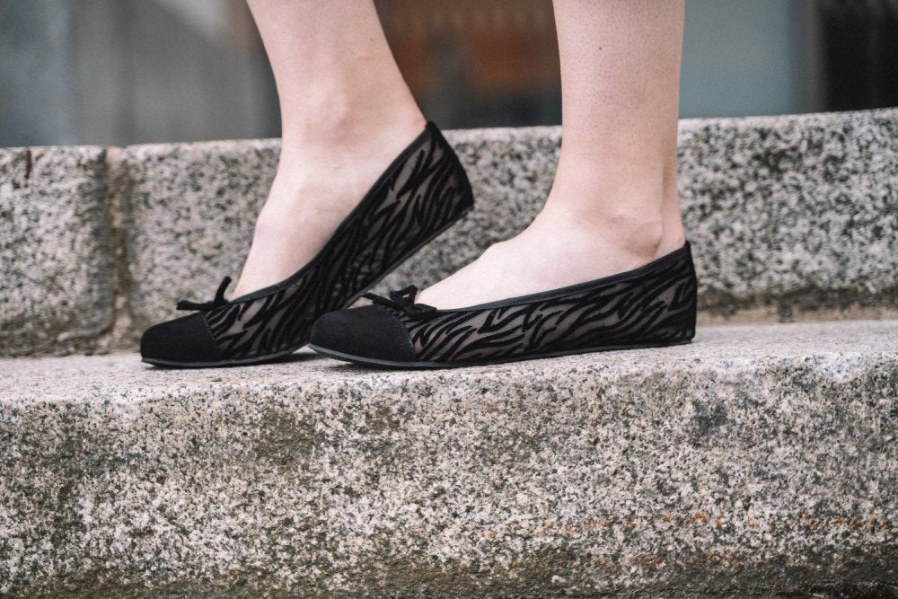 Nahaufnahme einer Person, die schwarze Ballerinas mit Zebrastreifen und ZAQQs TWIST Sling Black-Branding trägt und auf Steinstufen steht. Die Schuhe haben vorne ein Schleifendetail und eine flexible Sohle für zusätzlichen Komfort.