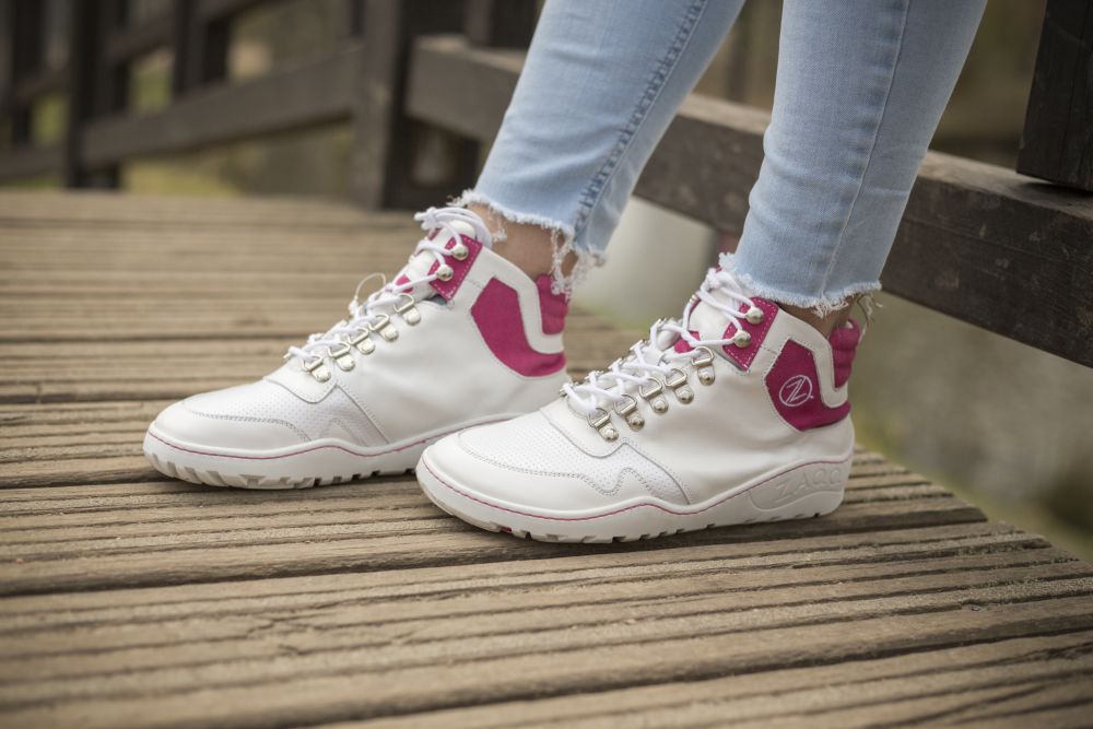 Eine Person, die ZAQQs VAQATION Pink Waterproof High-Top-Sneaker mit goldenen Ösen und einem markanten Logo trägt, steht auf einer Holzbrücke. Ihre hellblauen, an den Knöcheln ausgefransten Jeans verleihen dem farbenfrohen Schuhwerk eine lässige Note.