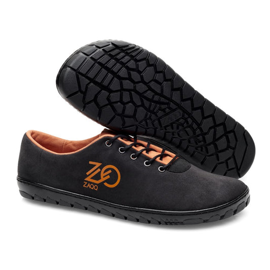 Die QARO Black Orange Schuhe von ZAQQ werden nebeneinander präsentiert, wobei einer aufrecht steht, während der andere seine flexible Kautschuksohle hervorhebt. Sie bestechen durch ihr stilvolles Design mit orangefarbenem Innenfutter und seitlichem Branding und bieten sowohl Eleganz als auch Komfort.