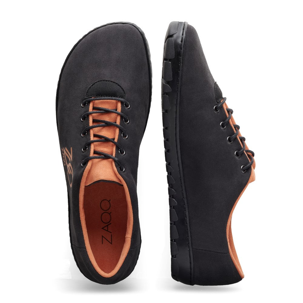 Ein Paar schwarze QARO Black Orange Sneaker von ZAQQ, mit minimalistischem Design mit hellbraunem Futter und Schnürsenkeln. Der linke Schuh ist von oben zu sehen und zeigt den Markennamen im Inneren dieses veganen Sneakers, während der rechte von der Seite zu sehen ist, um seine flexible Kautschuksohle hervorzuheben.