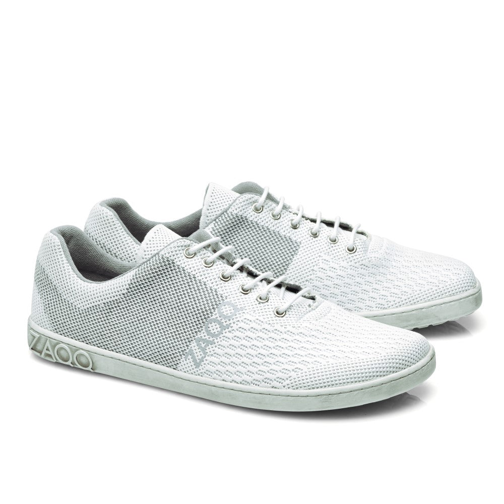 Wir präsentieren den QNIT White von ZAQQ, ein Paar hellgraue Low-Top-Sneaker mit atmungsaktiver Mesh-Textur und Schnürsenkeln. Dieser vegane Barfußschuh besticht durch ein elegantes Design mit dem Markenlogo an der Seite und der Sohle und ist damit perfekt für alle, die modernen Stil und Komfort schätzen.