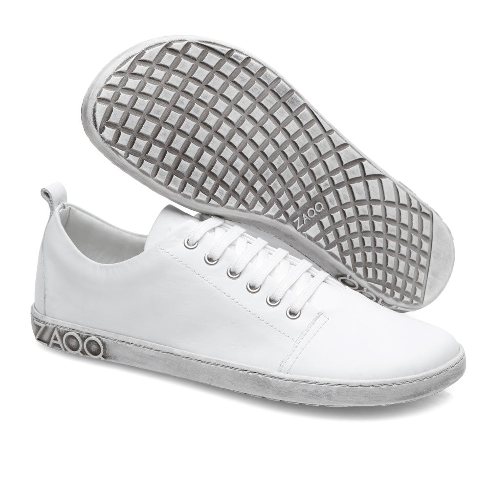 Ein Paar TAQQ Nappa White Sneaker von ZAQQ aus hochwertigem Nappaleder mit strukturiertem Muster und grauen Sohlen. Der linke Barfußschuh steht aufrecht und zeigt die Seitenansicht mit dem ZAQQ-Label auf der flexiblen Kautschuksohle, während der rechte Schuh geneigt ist, um das Design der unteren Sohle zu zeigen.